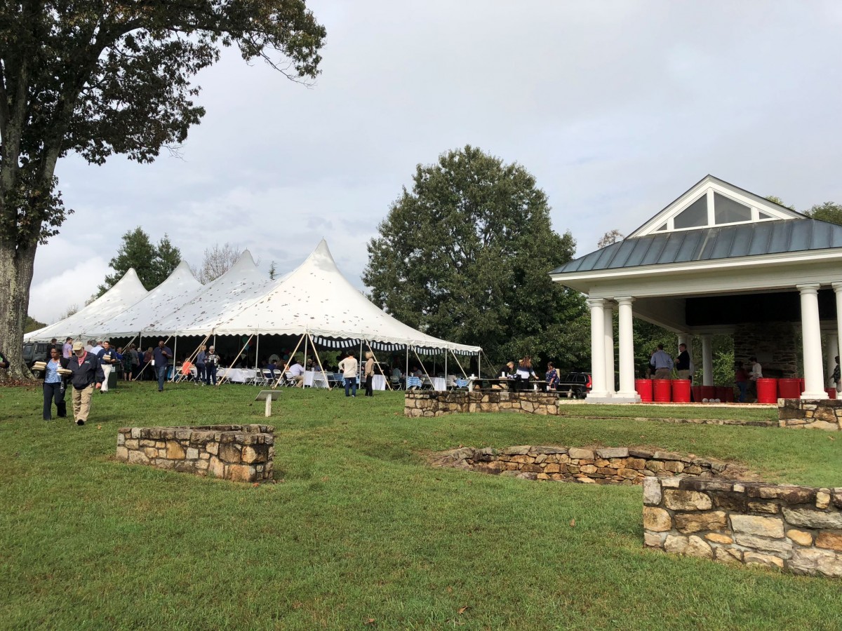 a festive white event tent sits on a green lawn while people eat and drink in early fall