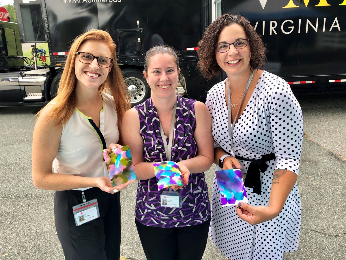 Three smiling women display small pieces of colorful artwork they created