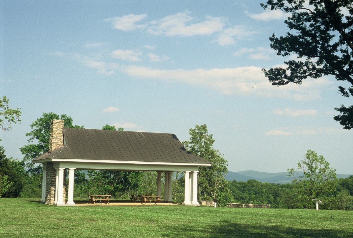 two wood picnic tables sit under a white pillared and stone pavilion